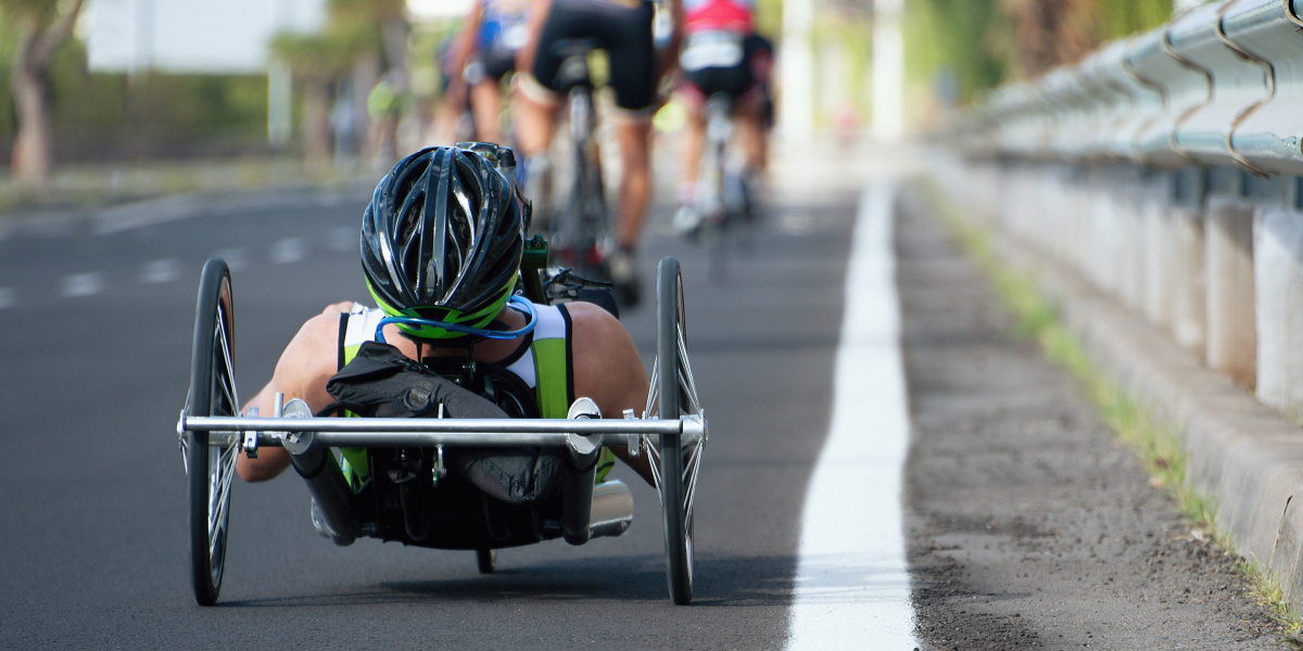 Mężczyzna na handbike’u jedzie po szosie w grupie kolarzy, widziany od tyłu podczas wyścigu na otwartej drodze.